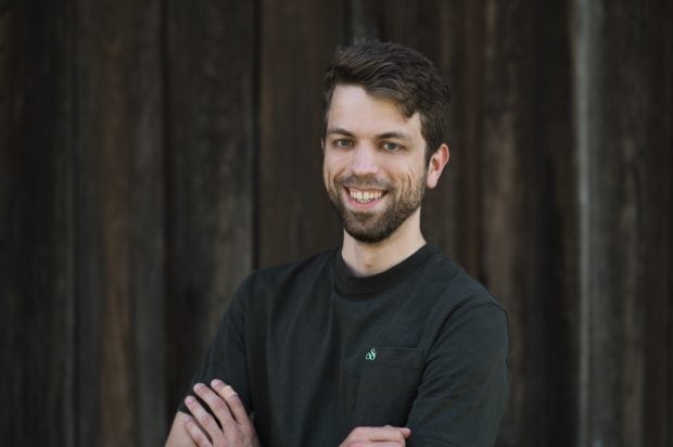 Ein junger Mann steht mit verschränkten Armen vor einer dunklen Holzwand. Er lächelt in die Kamera.