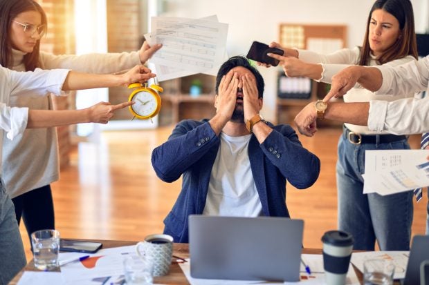 Mann am Schreibtisch wird von links und rechts mit Papieren, Weckern etc. bedrängt und ist sichtlich gestresst