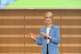 Lisa-Charlotte Wolter steht in einem blauem Blazer vor einem braunen Hintergrund und hält einen Vortrag.