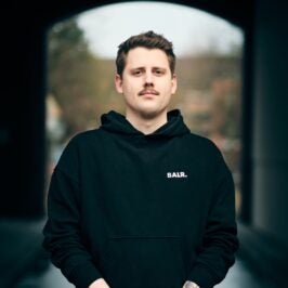 Nils Mertens steht in einem schwarzen Hoodie draußen, vor einem Durchgang.