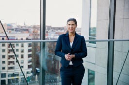 Katja Heintschel von Heinegg steht in einem dunkelblauen Anzug vor einem großen Fenster, hinter ihr ist eine Skyline zu sehen.