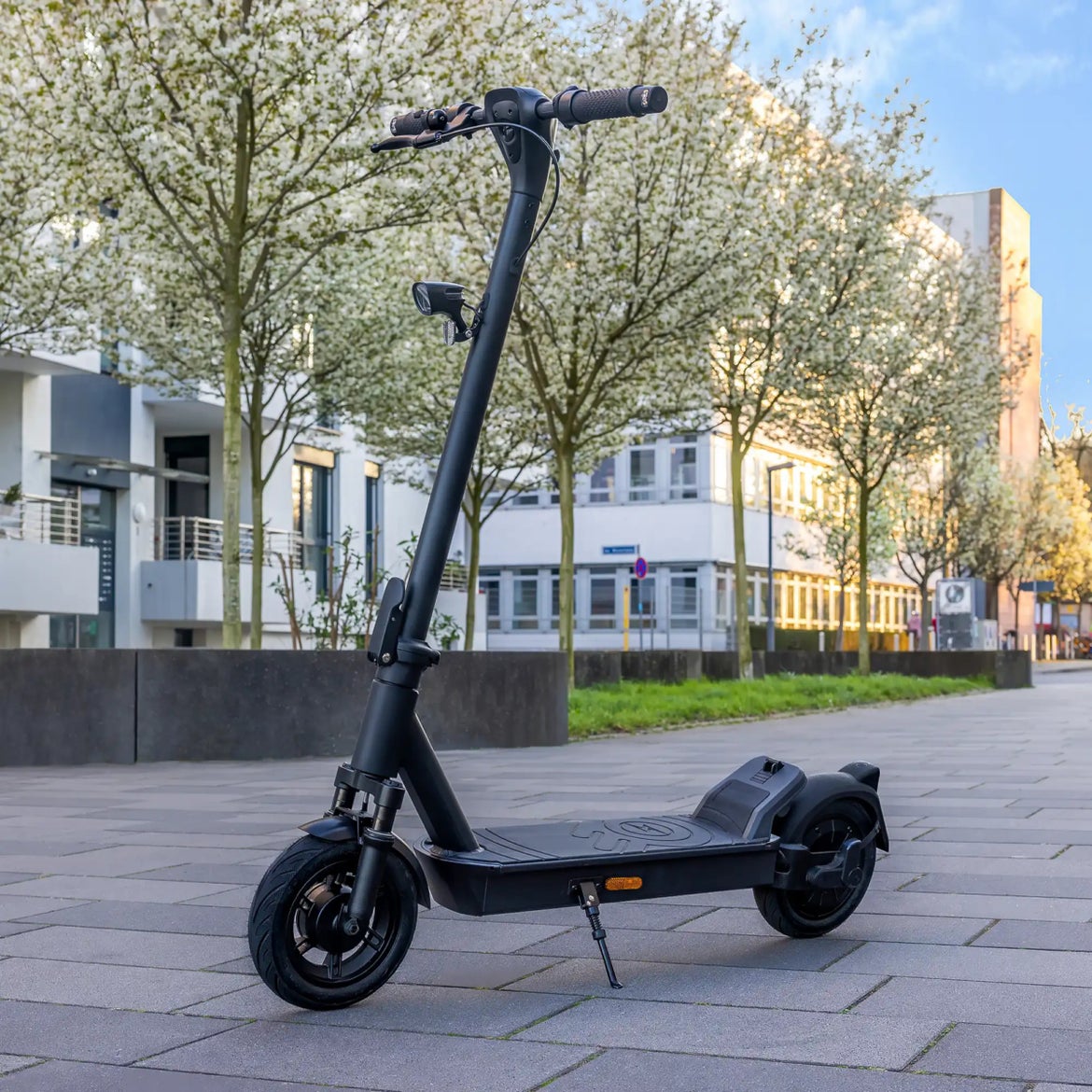 Schwarzer E-Scooter steht auf dem Seitenständer auf einem gepflasterten Platz vor blühenden Bäumen.