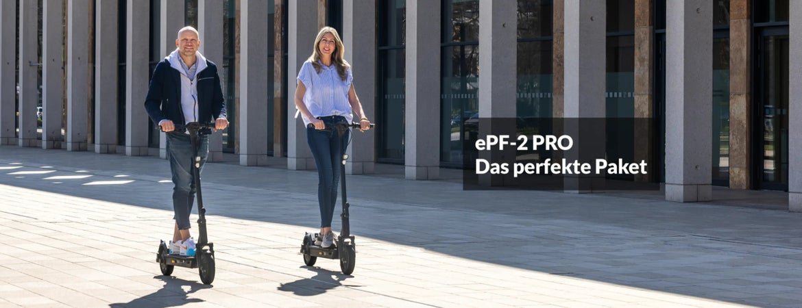 Zwei Personen fahren auf E-Scootern über einen gepflasterten Vorplatz vor einer Glasfassade mit Säulen.