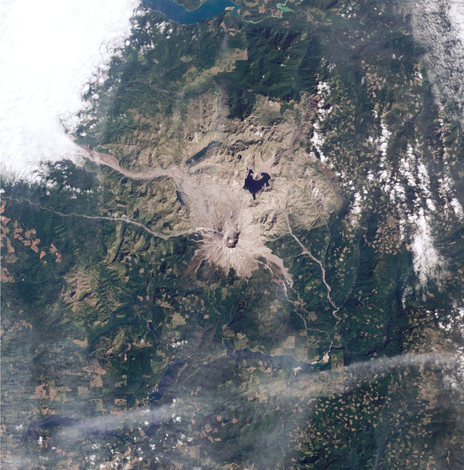 Eine Satelliten-Aufnahme zeigt den grau-braunen Mount St. Helens und die ihn umgebende Vulkanlandschaft, links im Bild ist eine große Wolke zu sehen.