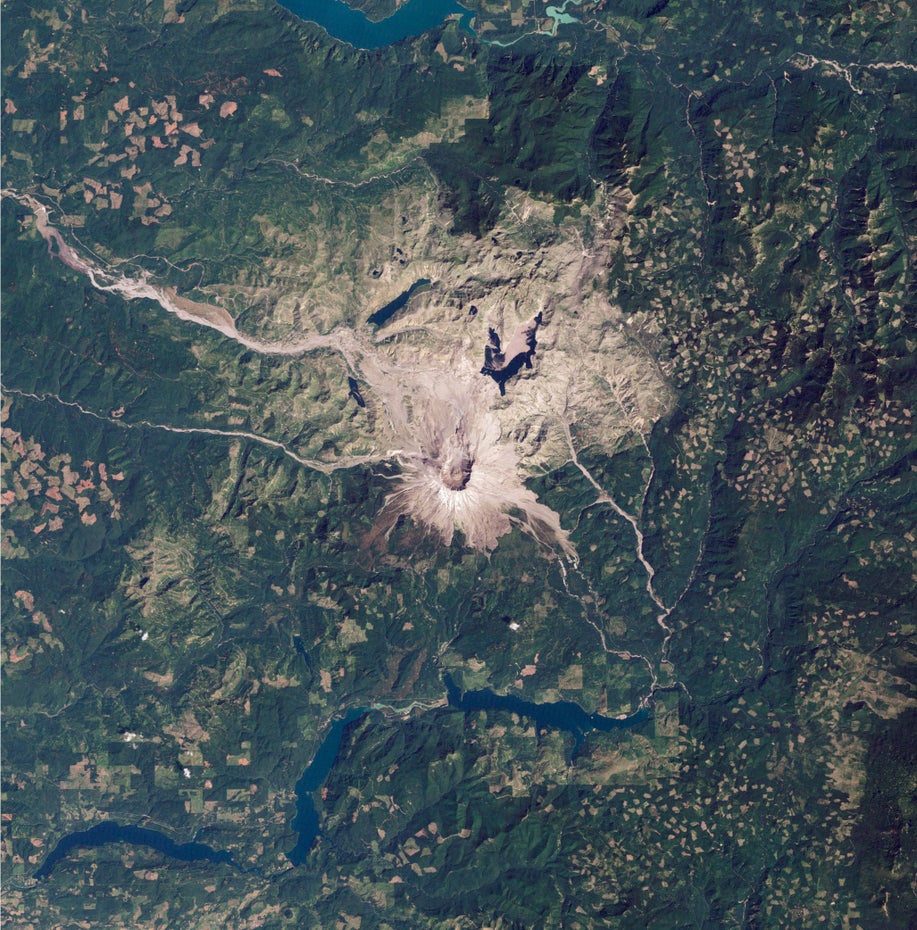 Der Krater des Mount St. Helens ist mittig im Bild zu sehen, ihn umgeben oberhalb eine grau-braune Vulkanlandschaft und unterhalb sowie links und rechts eine grüne Landschaft, in der auch Stauseen zu sehen sind.