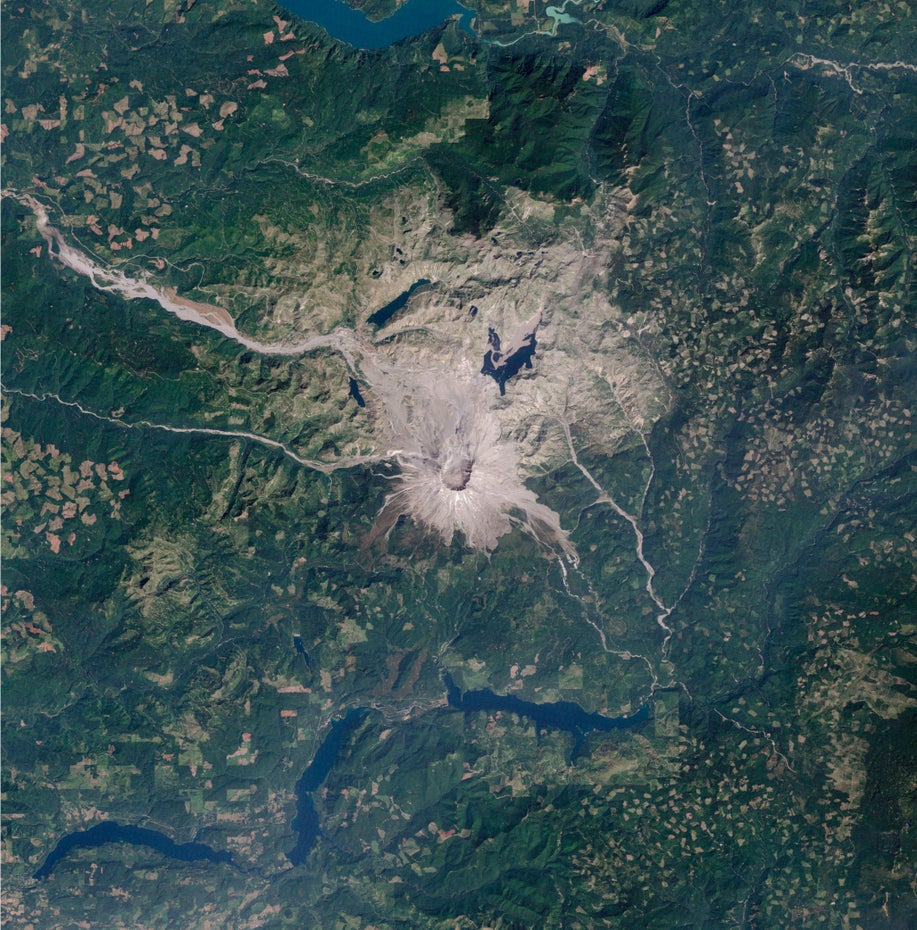 Der grau-braune Vulkan Mount St. Helens ist mittig im Bild zu sehen, ihn umgibt eine größtenteils grüne Landschaft.
