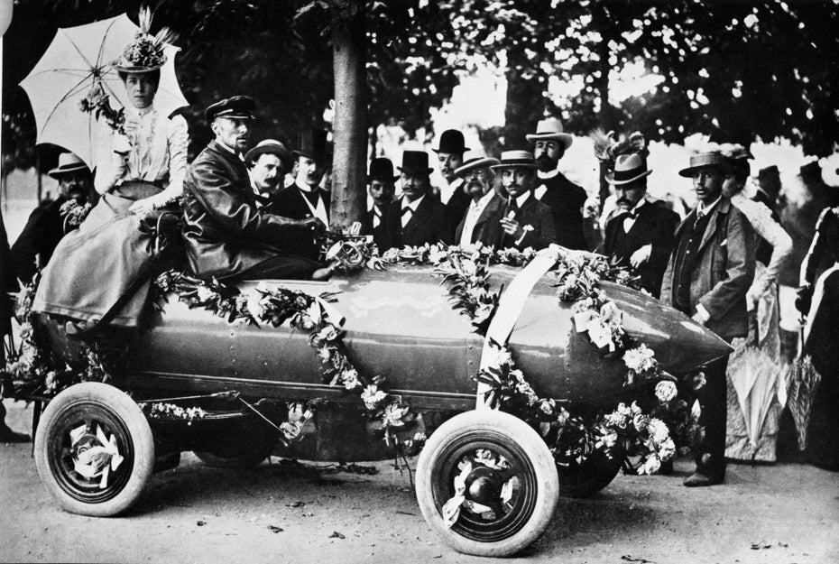 Fotoaufnahme in Schwarz-Weiß: Camille Jenatzy sitzt mit Mantel und Schirmmütze bekleidet in einem länglichen Auto, das mit Blumengirlanden geschmückt ist.