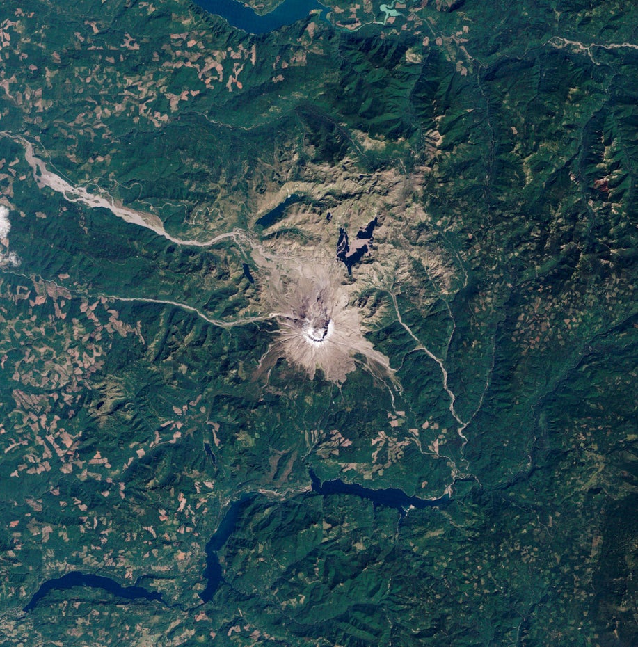Der Krater des Mount St. Helens ist mit weißem Schnee bedeckt, der Vulkan und seine nähere Umgebung liegen grau-braun gefärbt in einer grünen Landschaft.