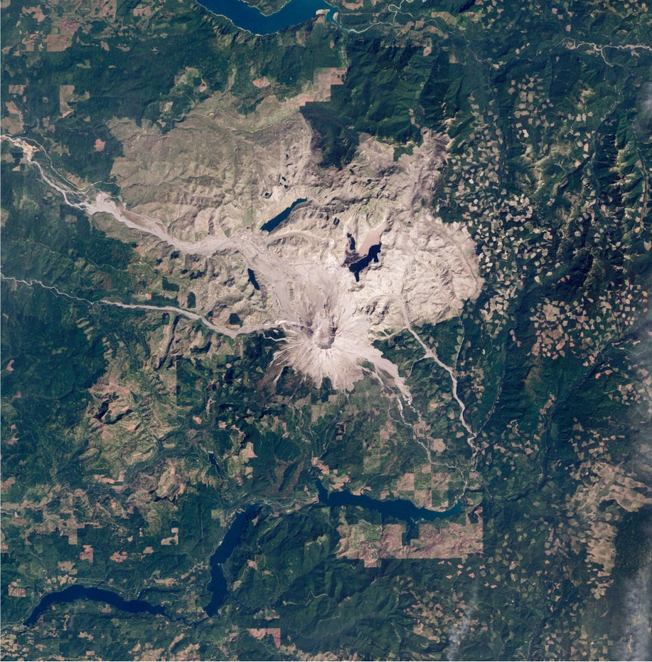 Mittig im Bild ist in grau-braun der Vulkan und die Landschaft um ihn herum zu sehen, darum ist grüne Landschaft sichtbar.