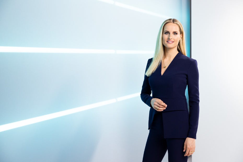 Janna Ensthaler steht in einem schwarzen Hosenanzug vor einer blauen Wand.