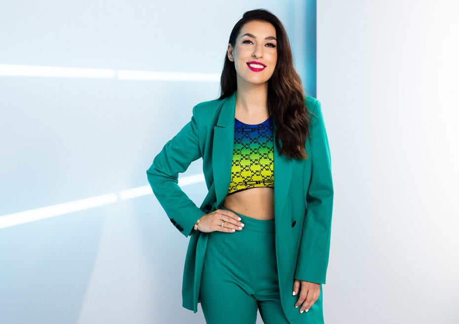 Tijen Onaran steht in einem grünen Hosenanzug vor einer hellblauen Wand.