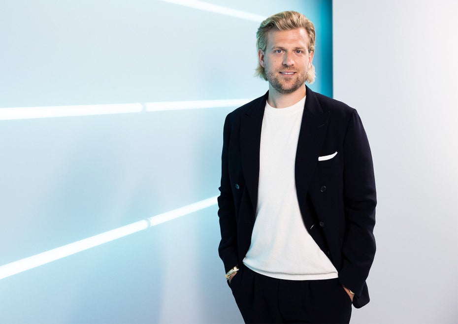 Tillman Schulz steht in einem schwarzen Anzug vor einer hellblauen Wand.