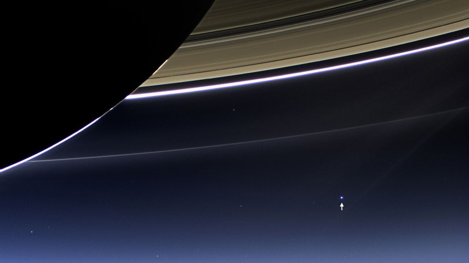 Die Erde ist ein kleiner blauer Fleck in der Ferne, im Vordergrund ist der Saturn mit seinen mächtigen Ringen zu sehen.