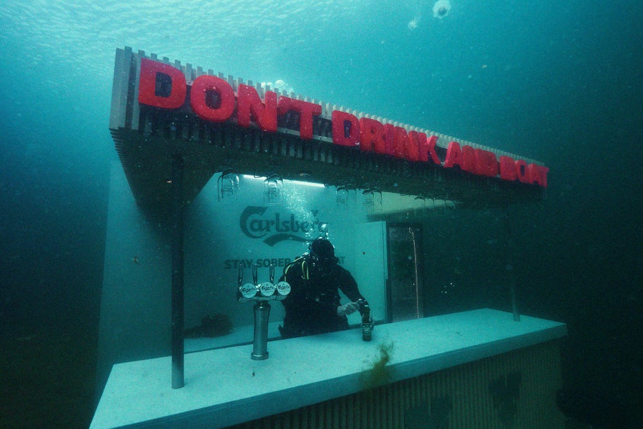 Auf einer Unterwasserbar steht der rote Slogan „Don't drink and drive”, an der Bar ist ein Taucher.