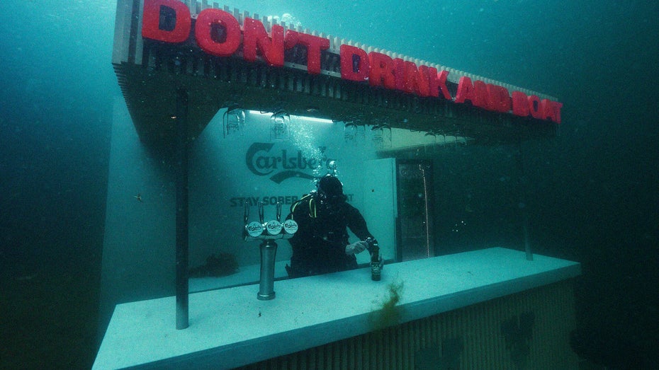 Auf einer Unterwasserbar steht der rote Slogan „Don't drink and drive”, an der Bar ist ein Taucher.